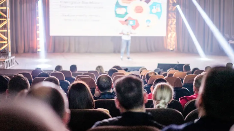 Audience in a conference hall watching a speaker on a lit stage