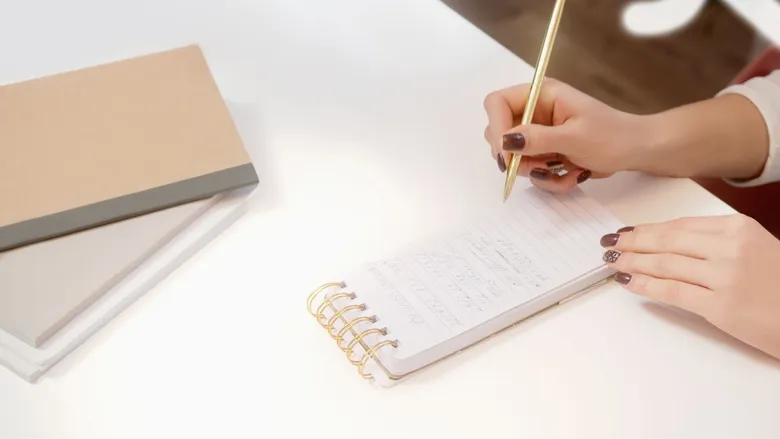 Close-up of hands writing notes in a spiral notebook on a white desk